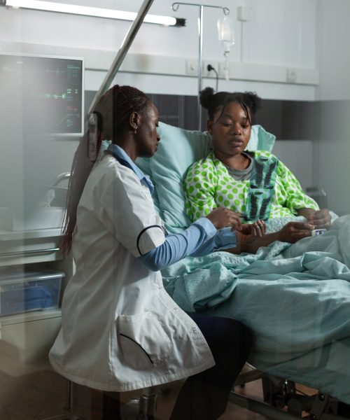 Doctor of african american ethnicity holding radiography in hospital ward. Afro woman looking at x ray with young patient for treatment recovery. Black girl sitting in bed talking to medic