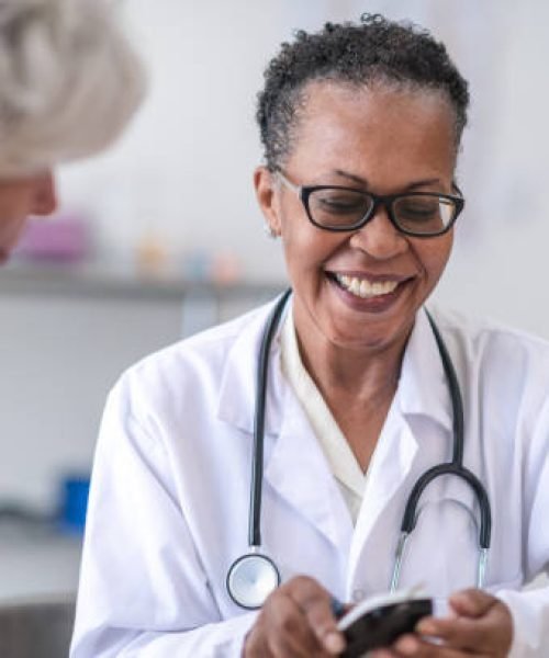 A friendly black female doctor is showing a senior women how to check her blood pressure because of diabetes. The doctor is wearing a white doctors jacket and a stethoscope around her neck. They are in a clinical setting.