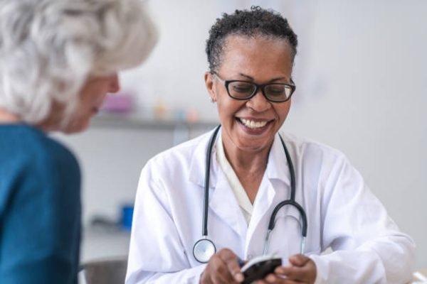 A friendly black female doctor is showing a senior women how to check her blood pressure because of diabetes. The doctor is wearing a white doctors jacket and a stethoscope around her neck. They are in a clinical setting.