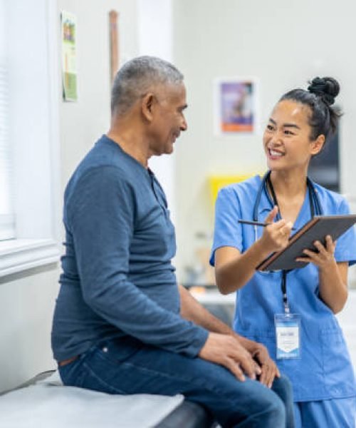 A senior gentleman of Hispanic decent, sits up on an exam table during a routine check-up at the doctors.  A nurse, who is wearing blue scrubs, is standing in front of the gentleman as she reviews some recent test results with the patient on a tablet.