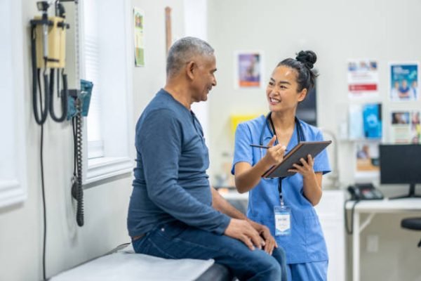 A senior gentleman of Hispanic decent, sits up on an exam table during a routine check-up at the doctors.  A nurse, who is wearing blue scrubs, is standing in front of the gentleman as she reviews some recent test results with the patient on a tablet.
