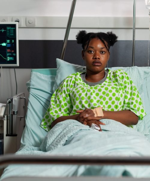 Portrait of young patient lying in bed during clinical examination recovering after medical surgery in hospital ward. Sick woman looking into camera waiting for healthcare treatment