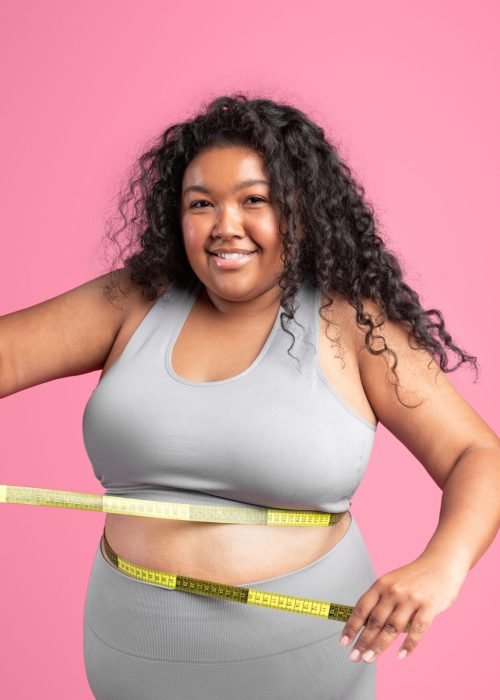 Smiling black overweight woman in sportswear checking waist with measuring tape, isolated on pink studio background. Sports, weight loss result and body care, diet, ad and offer