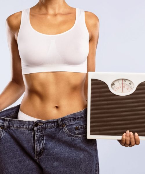 Closeup cropped view of unrecognizable woman in oversized jeans holding scales, showing results of weight loss program or liposuction. Lady achieving success in slimming, blue studio, banner, panorama