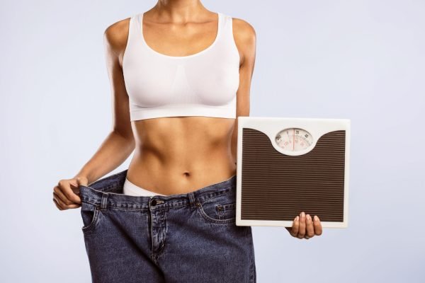 Closeup cropped view of unrecognizable woman in oversized jeans holding scales, showing results of weight loss program or liposuction. Lady achieving success in slimming, blue studio, banner, panorama