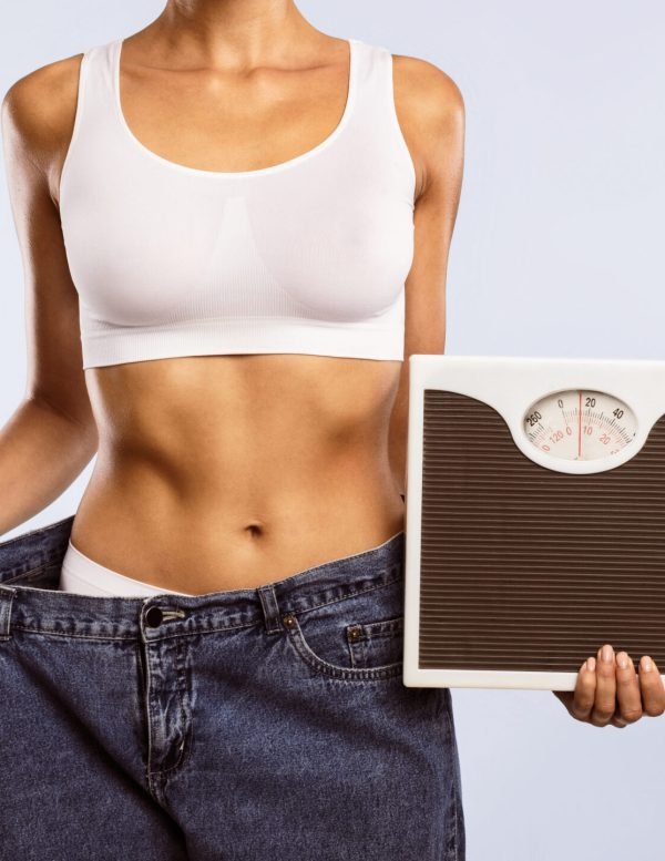 Closeup cropped view of unrecognizable woman in oversized jeans holding scales, showing results of weight loss program or liposuction. Lady achieving success in slimming, blue studio, banner, panorama